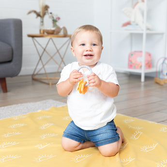 A toddler kneels on a Bumkins Reversible Roll & Go Blanket, Desert Boho, holding teething rings in a bright living room.
