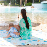 By the pool, a woman and baby sit on a Bumkins Reversible Roll & Go Blanket, Rolling with the Waves, as another child swims nearby.