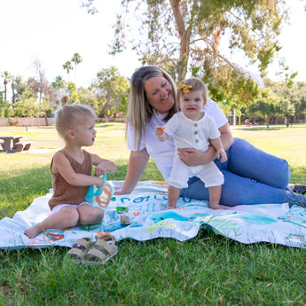 A woman and her kids sit on a Bumkins Reversible Roll & Go Blanket, Rolling with the Waves on the grass, enjoying a day at the park.