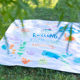 Bumkins Reversible Roll & Go Blanket, Rolling with the Waves lays on lush green grass, with leaves blurred in the foreground.