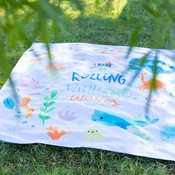Bumkins Reversible Roll & Go Blanket, Rolling with the Waves lays on lush green grass, with leaves blurred in the foreground.