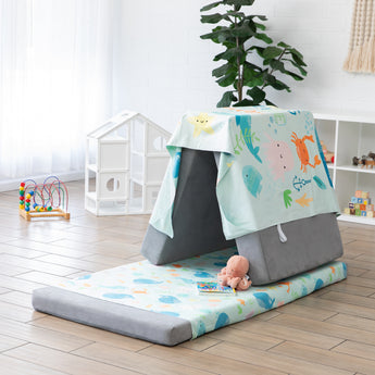 A play area with a gray cushion tent covered in a Bumkins Reversible Roll & Go Blanket, Rolling with the Waves, stuffed animal, and playhouse.