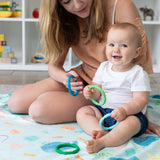 A baby sits on a Bumkins Reversible Roll & Go Blanket, Rolling with the Waves, holding colorful teething rings as an adult watches.