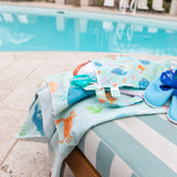 A lounge chair with a Bumkins Reversible Roll & Go Blanket, Rolling with the Waves, a bag, goggles, and blue sandals by a turquoise pool.