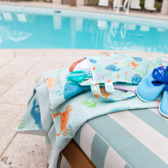 A lounge chair with a Bumkins Reversible Roll & Go Blanket, Rolling with the Waves, a bag, goggles, and blue sandals by a turquoise pool.