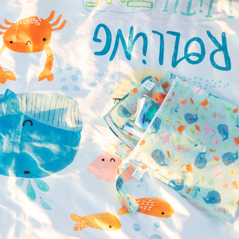 A bag with snacks and toys sits on a Bumkins Reversible Roll & Go Blanket, Rolling with the Waves. 