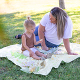 A woman and child sit on a Bumkins Reversible Roll & Go Blanket, Happy Camper on the grass, sharing a book while the child holds a card.
