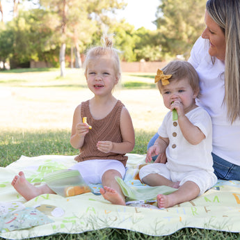 Two kids savor snacks on a sunny park day with a woman beside them on a Bumkins Reversible Roll & Go Blanket, Happy Camper.