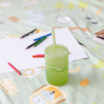 Bumkins Silicone Straw Cup with Lid: Sage  sits on a patterned blanket among paper and colored pencils.