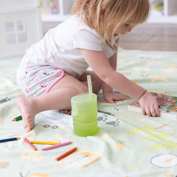 A girl on a patterned blanket reaches for a book, with colored pencils and a Bumkins Silicone Straw Cup with Lid: Sage close by.