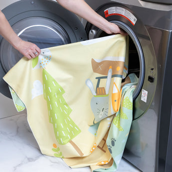 A person places a Bumkins Reversible Roll & Go Blanket, Happy Camper into an open front-loading washing machine.