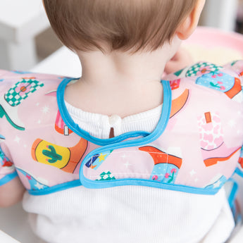 Close-up a child wearing a Bumkins Ruffle Apron Bib: Boots, with focus on the adjustable hook-and-loop closure.