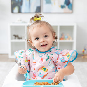 A toddler sits in a high chair, wearing a Bumkins Ruffle Apron Bib: Boots and holding a fork and spoon over a blue plate of waffles.