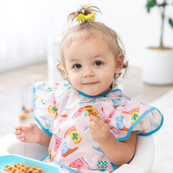 A toddler with a ponytail enjoys waffles in a high chair, wearing a Bumkins Ruffle Apron Bib: Boots in a bright room.