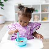 A young girl in a bib from Bumkins Snack In Style 3-Piece Set: Unicorns and Rainbows eats from a blue bowl in a high chair.