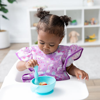A young girl in a bib from Bumkins Snack In Style 3-Piece Set: Unicorns and Rainbows eats from a blue bowl in a high chair.