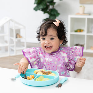 A young girl in a bib from Bumkins Snack In Style 3-Piece Set: Unicorns and Rainbows eats from a blue divided plate at a white table.