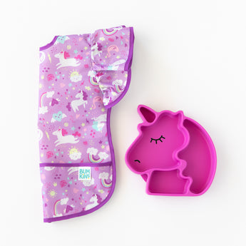 Bumkins Ruffle Apron Bib: Unicorns lays folded lengthwise next to a pink unicorn-shaped divided plate on a white background.