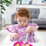 A toddler in a high chair, wearing a Bumkins Ruffle Apron Bib: Unicorns, holds a blue spoon over a pink divided plate of food.