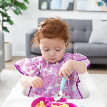 A toddler in a high chair, wearing a Bumkins Ruffle Apron Bib: Unicorns, holds a blue spoon over a pink divided plate of food.