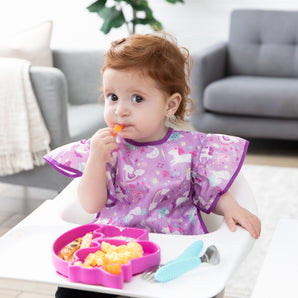 A toddler in a high chair eats snacks from a pink unicorn-shaped plate, wearing a Bumkins Ruffle Apron Bib: Unicorns in a cozy living room.