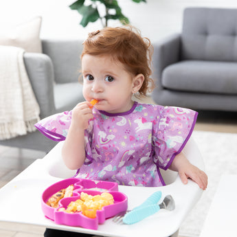 A toddler in a high chair eats snacks from a pink unicorn-shaped plate, wearing a Bumkins Ruffle Apron Bib: Unicorns in a cozy living room.