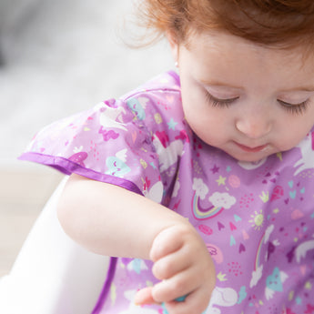 Close-up of a toddler looking down in a high chair, wearing a Bumkins Ruffle Apron Bib: Unicorns.