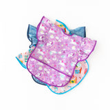 Bumkins Ruffle Apron Bib: Unicorns lays on top of a stack of similar bibs on a white background.