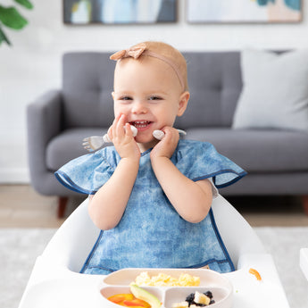 A smiling toddler in a Bumkins Ruffle Apron Bib: Crushed Denim sits in a high chair, holding utensils by their cheeks with a plate nearby.
