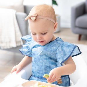 In a bright room, a toddler enjoys food in a high chair, wearing a Bumkins Ruffle Apron Bib: Crushed Denim and a bow headband.