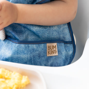 Close-up of a child in a high chair wearing a Bumkins Ruffle Apron Bib: Crushed Denim, with focus on the pocket.
