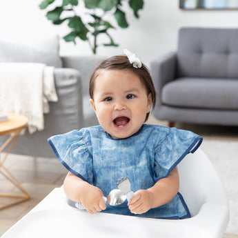 A smiling baby in a bow and a Bumkins Ruffle Apron Bib: Crushed Denim sits in a high chair in a cozy living room, holding utensils.