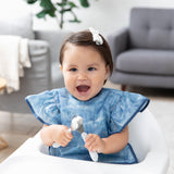 A girl in a blue bib beams in a high chair, holding Bumkins Spoon + Fork: Sand in a cozy living room.