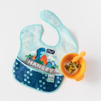 A bib from Bumkins SuperBib® and Sleeved Bib 3-Piece Set: Hangry, Dinosaurs, Blue Tropics rests next to an orange bowl & spoon with yogurt.