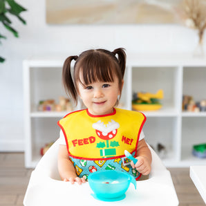 A girl in pigtails wearing a Bumkins SuperBib® 2 Pack: Super Mario™ Expressions smiles in a high chair with a blue bowl on the tray.