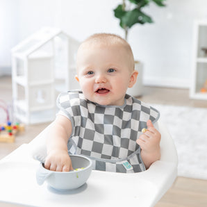 A baby in a high chair, smiling in a Bumkins SuperBib® 3 Pack: Palms + Check, enjoys eating from a bowl with a small playhouse nearby.