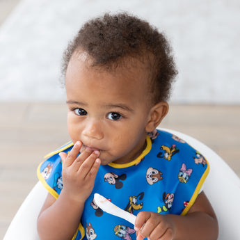 A young boy stares curiously, wearing a bib from Bumkins On-The-Go Mealtime 7-Piece Set: Mickey & Friends MVP.