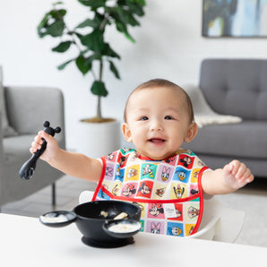 A baby smiles in a colorful Bumkins SuperBib® 3 Pack: Mickey & Friends MVP, holding a spoon near a Mickey Mouse-shaped bowl with yogurt.