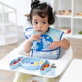 A girl wearing a Bumkins SuperBib® 3 Pack: Stitch Star-ring Up Trouble eats from a matching plate in a high chair in a bright playroom.