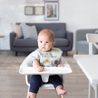 A baby sits in a high chair in a cozy living room, wearing a bib from Bumkins On-The-Go Mealtime 7-Piece Set: Pooh Bear + Friends.