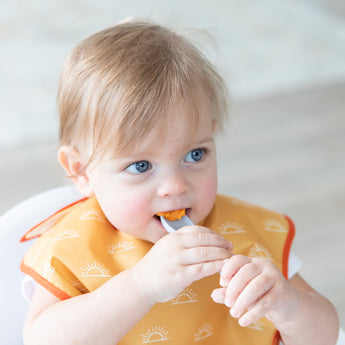 A boy in a yellow bib eats mashed sweet potatoes with a Bumkins Silicone Dipping Spoons 3 Pack: Taffy.