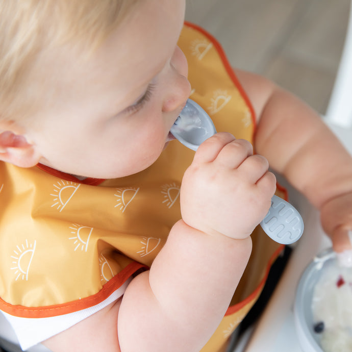 Close-up of a blonde child in a yellow bib practicing self-feeding with yogurt on a gray spoon. 