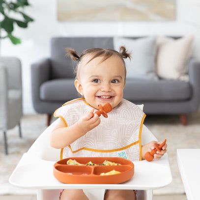 A smiling toddler with pigtails sits in a high chair, wearing a bib. They are holding a spoon and a fork with food on the tray in front of them. A couch and a plant are in the blurred background.