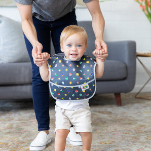 A smiling child wears a Bumkins SuperBib® 3 Pack: Super Mario™ Power-Up while and adult stands behind them, holding their hands.