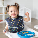 A smiling girl wearing a Bumkins SuperBib® 3 Pack: Super Mario™ Power-Up sits in a high chair, eating fruit & yogurt from a blue plate.