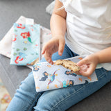 Close-up of a child holding a cookie with bags from Bumkins Snack In Character 4-Piece Set: Disney Princess Magic Tiana on their lap.