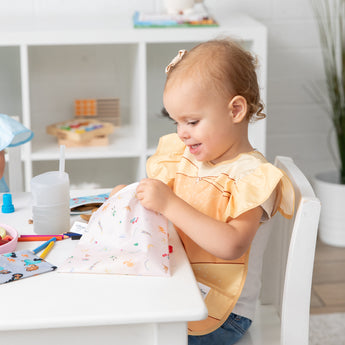 A smiling girl sits at a table in a playroom with Bumkins Snack In Character 4-Piece Set: Disney Princess Magic Belle.