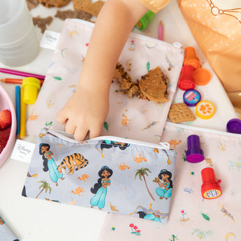 A child reaches for bags from Bumkins Snack In Character 4-Piece Set: Disney Princess Magic Belle, surrounded by snacks and art supplies.