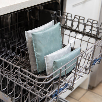 Four Bumkins Reusable Snack Bags in Desert Night are placed upright on the bottom rack of an open kitchen dishwasher.