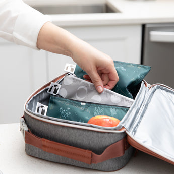 A hand places Roamio by Bumkins Reusable Snack Bag 4 Pack: Desert Night  in a lunch bag on a kitchen counter.